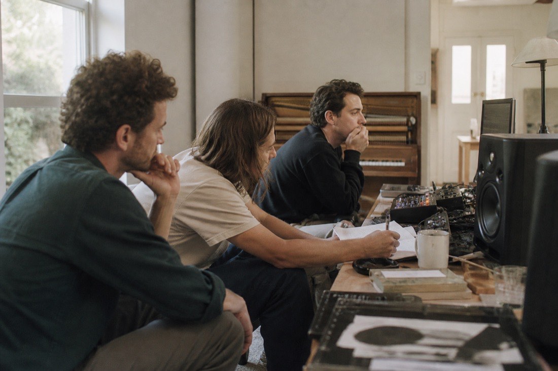 Three musicians listening to a track at studio monitors in a daylit room, one taking notes